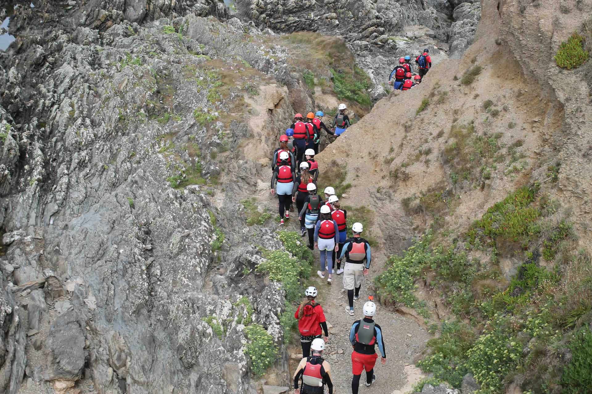 Group of people wearing helmets and life jackets walking along a narrow rocky path beside cliffs.