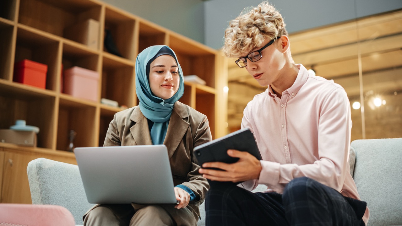 Young woman wearing a blue hijab and brown jacket working on a laptop with a young man in glasses and a pink shirt holding a tablet, seated in a modern office.