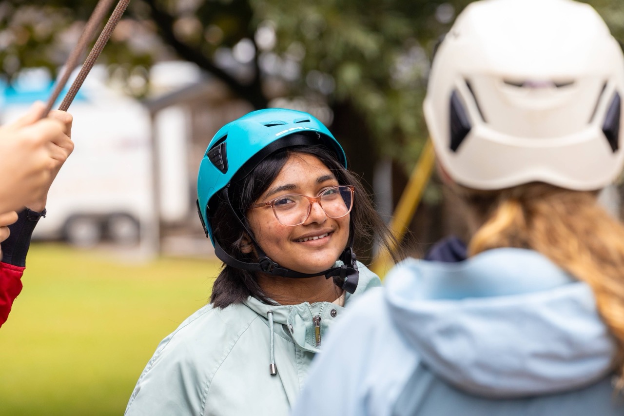 Smiling young woman wearing a teal helmet and glasses, outdoors in a park setting, interacting with another person in a white helmet.