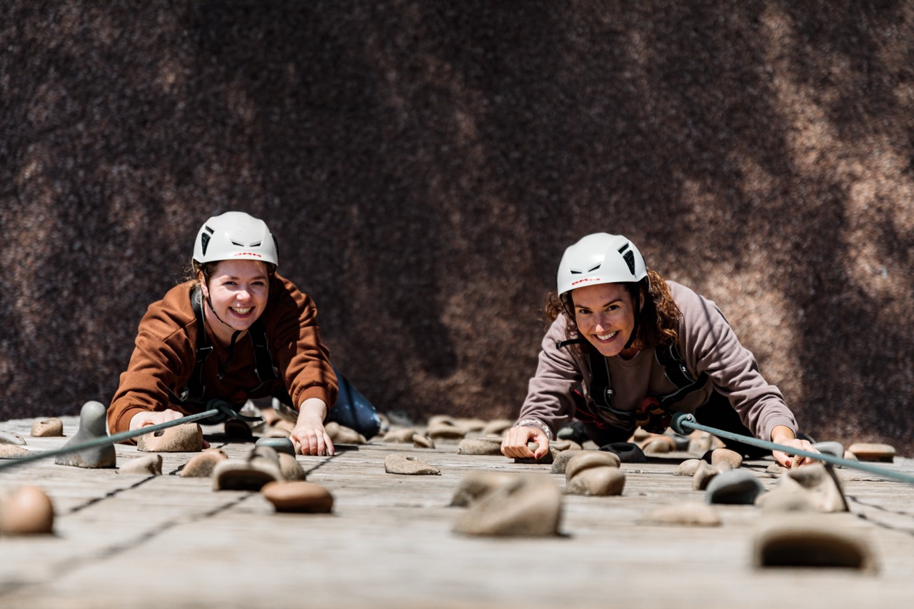 Two women wearing helmets and harnesses smiling as they climb an indoor rock climbing wall with handholds and footholds.