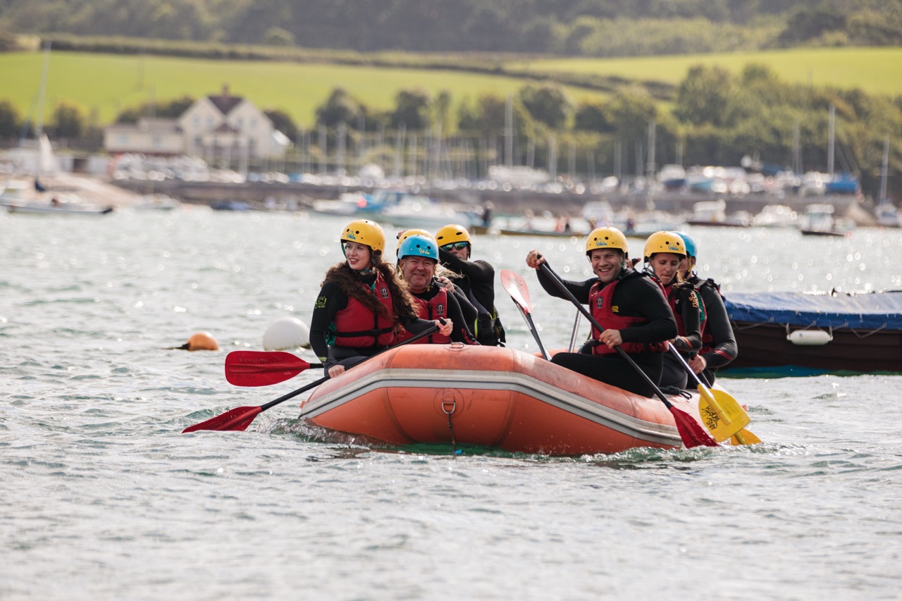 Group of six people wearing helmets and life jackets paddling an orange inflatable raft on a lake.