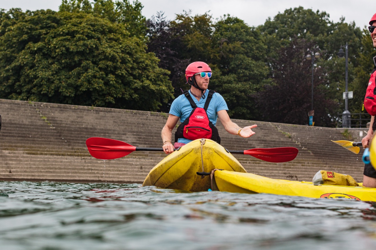 Man wearing a red helmet and life vest paddling a yellow kayak on water near a concrete embankment.