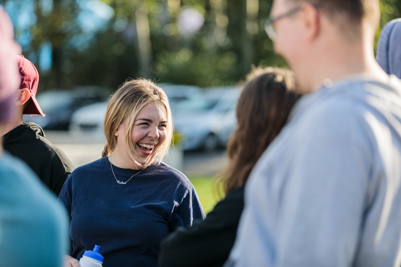 Smiling young woman wearing a blue shirt and a necklace with the name Rachael, talking outdoors with a group of people.
