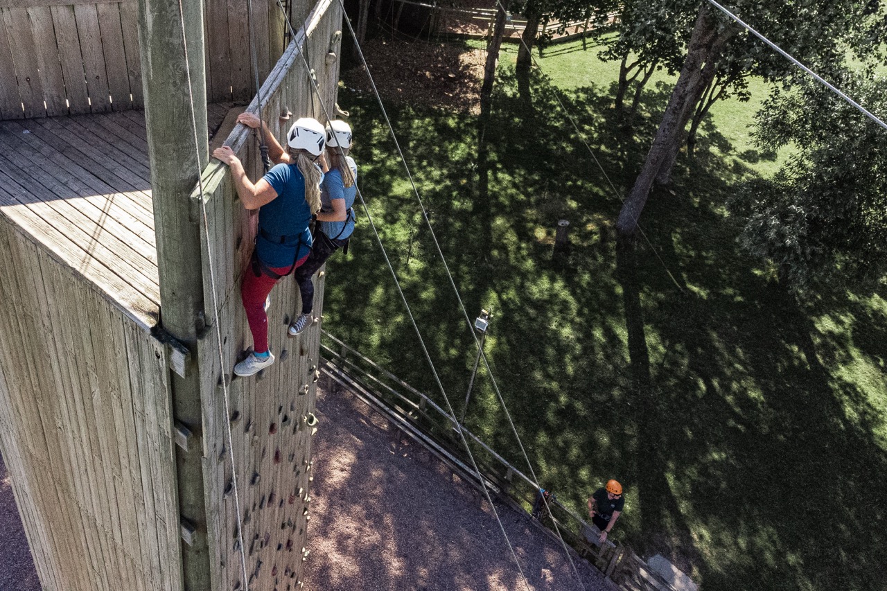 Two people wearing helmets climbing a tall outdoor wooden climbing wall with a person in an orange helmet on the ground below.