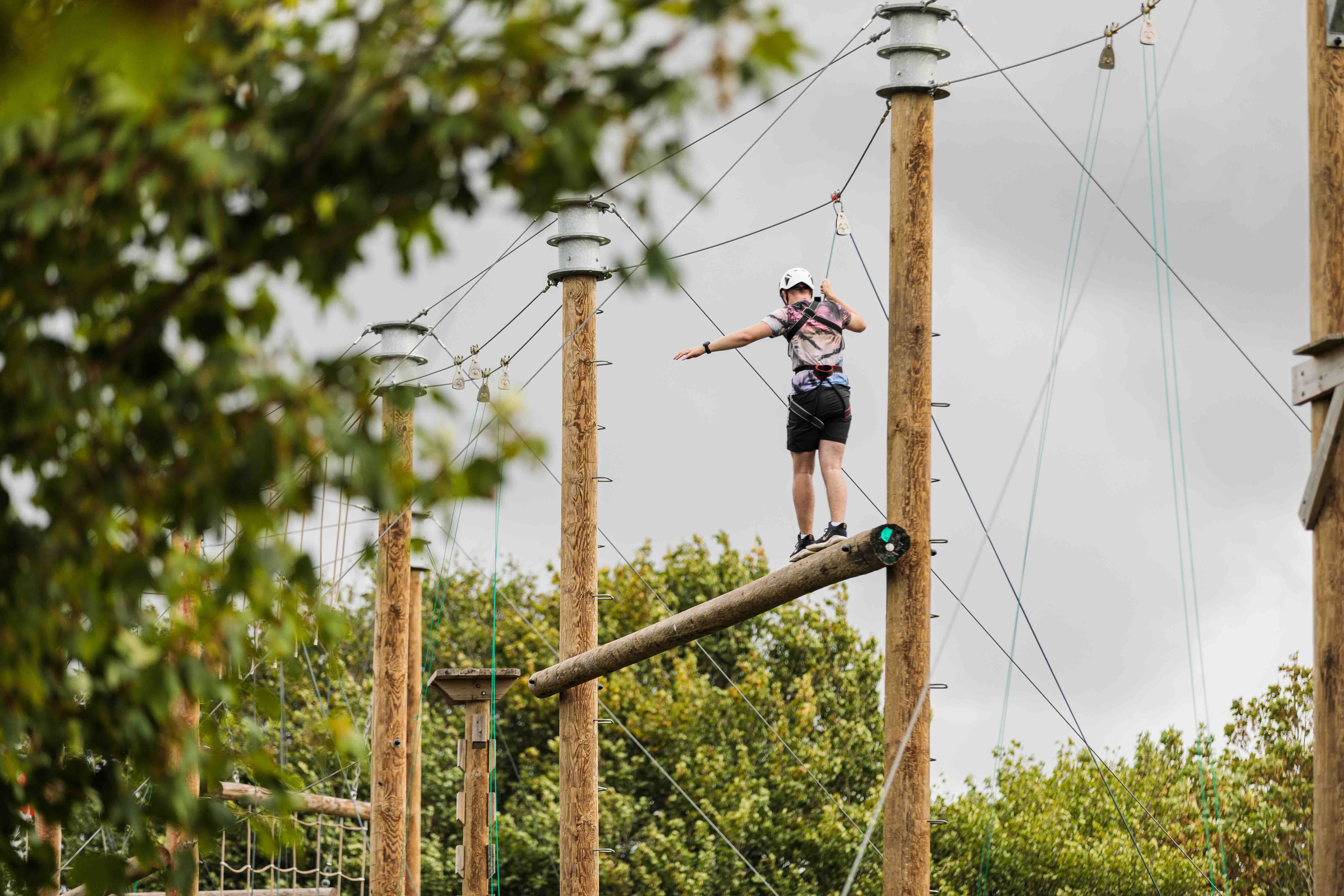 Person wearing helmet and harness walking on an elevated log in an outdoor ropes course.