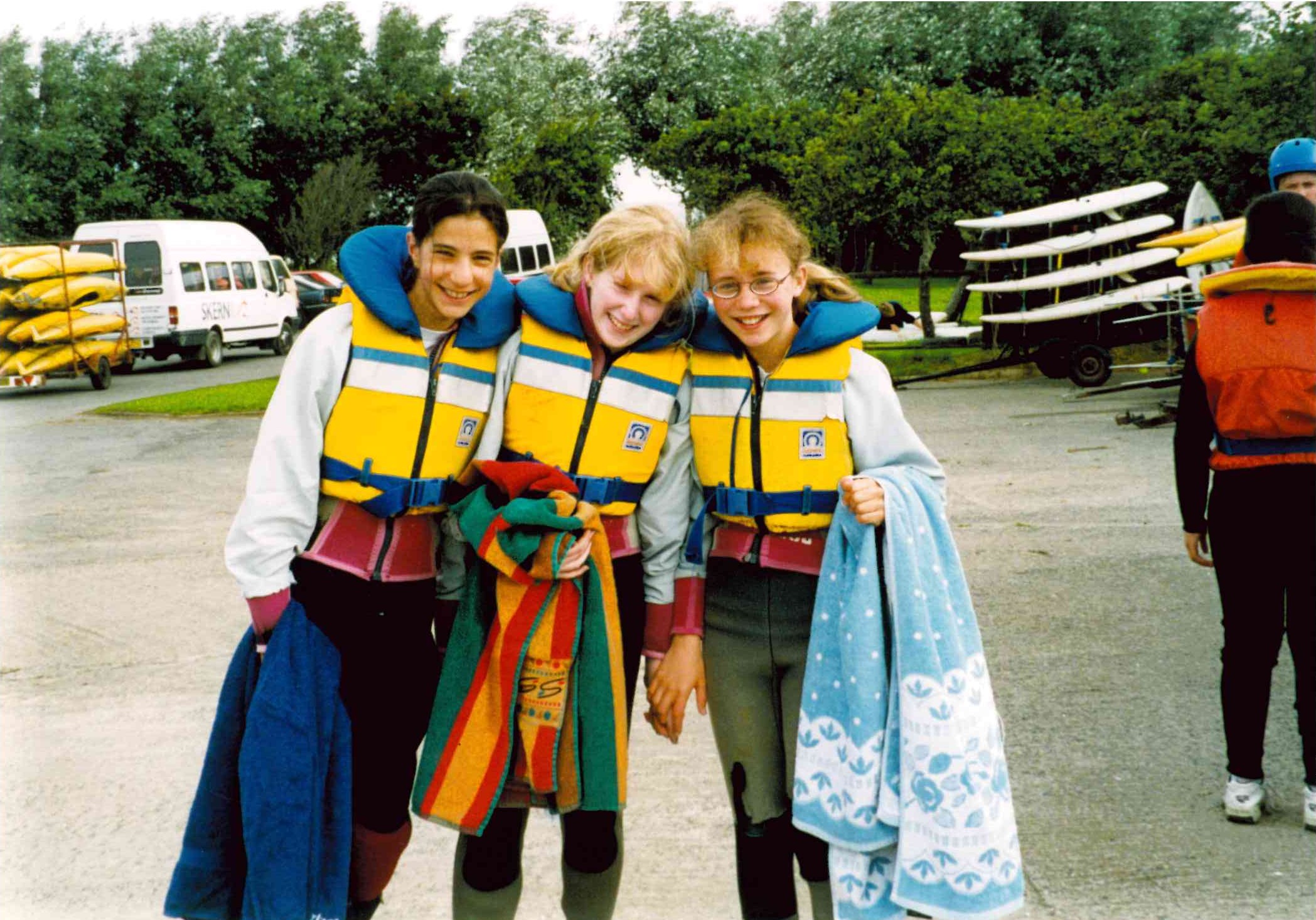 Three smiling girls in wetsuits and yellow life jackets holding towels by parked kayaks and vans.