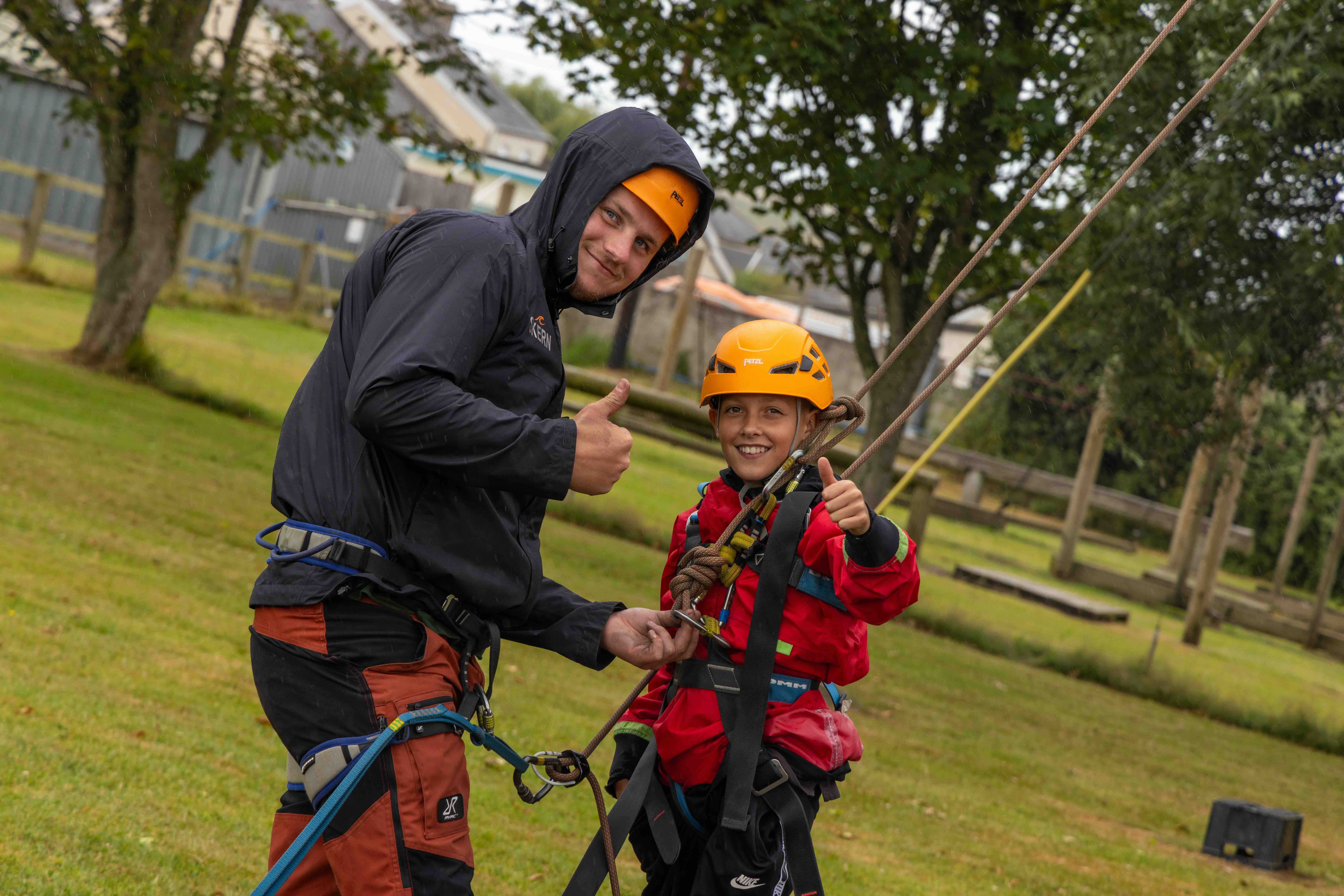 Man in black jacket and child in red jacket with helmets smiling and giving thumbs up during climbing activity.