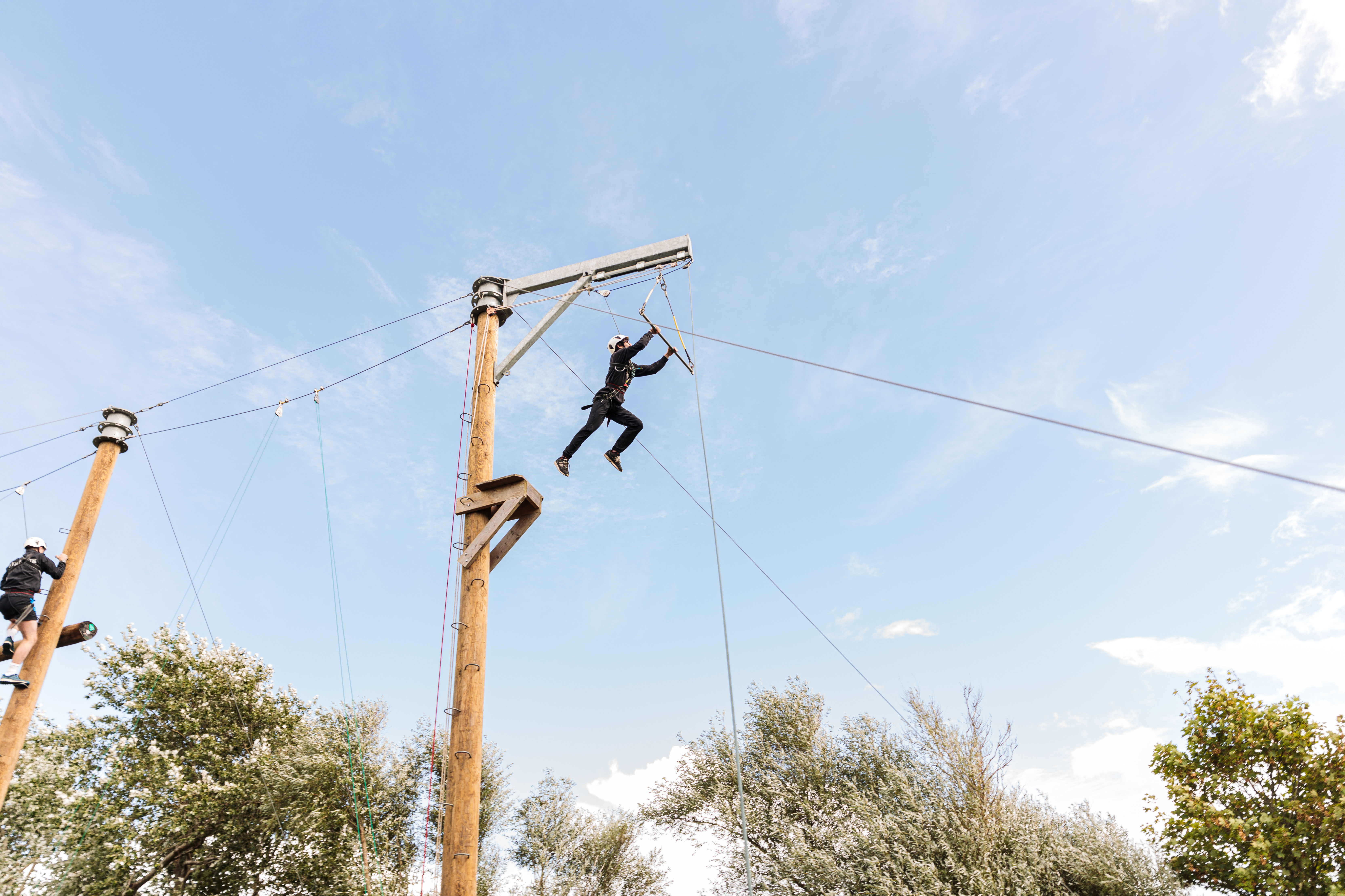 Person in safety gear jumping from platform to trapeze on outdoor ropes course with blue sky background.