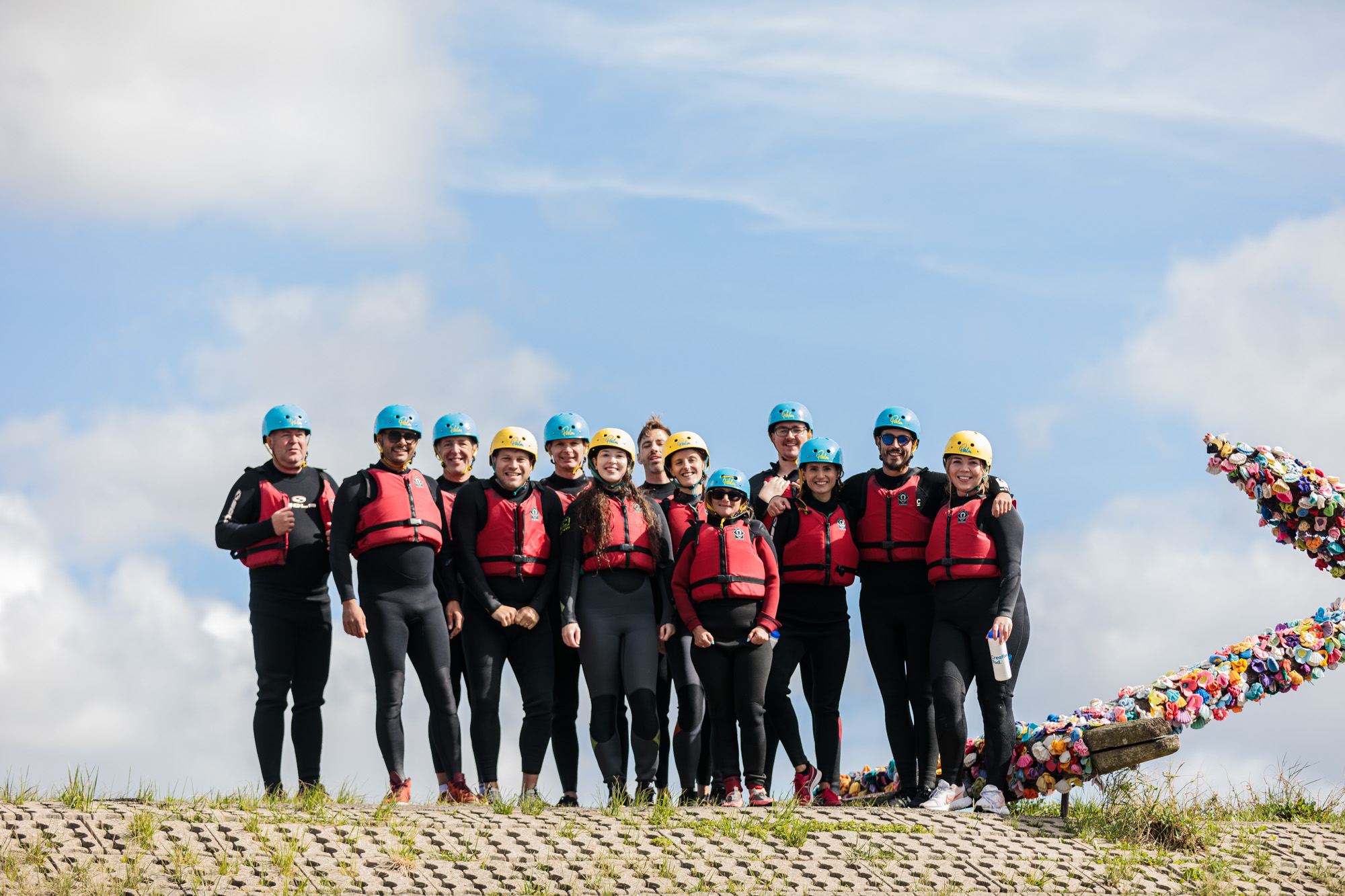 Group of people in wetsuits, red life jackets, and helmets standing outdoors under a blue sky.