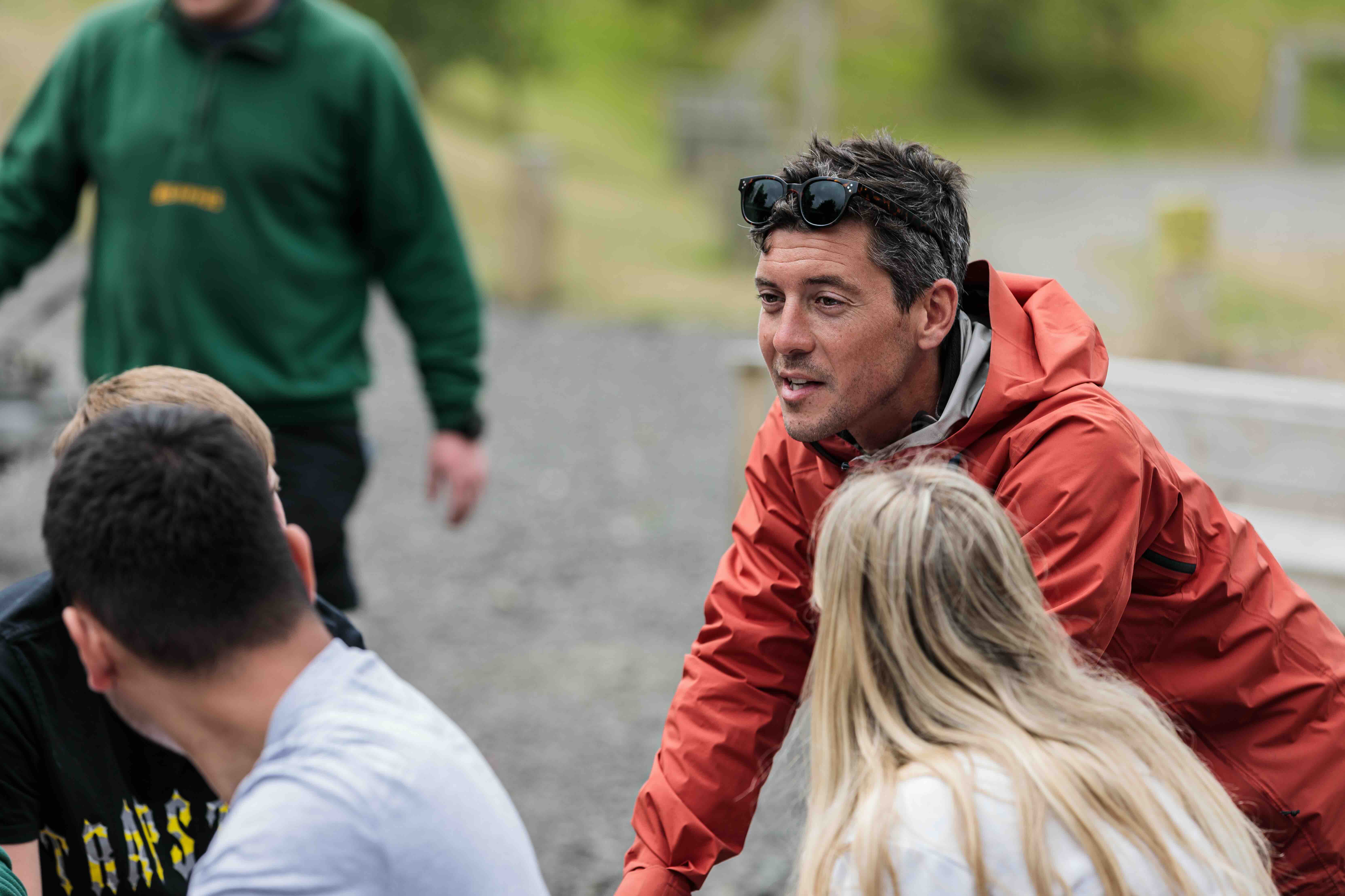 Man in orange jacket with sunglasses on head talking to a group outdoors on a gravel path.