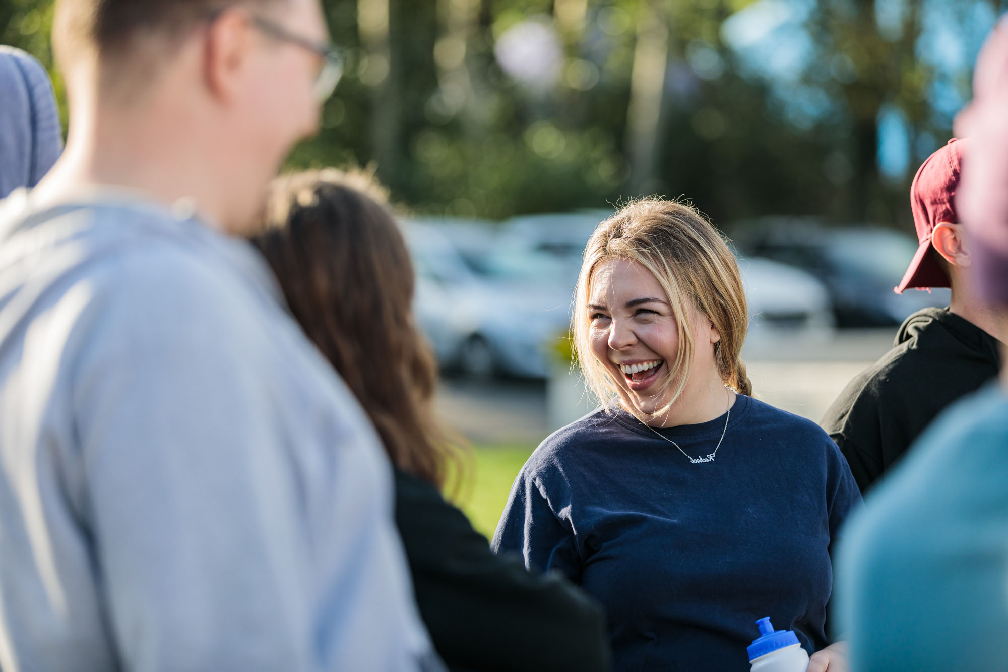 Smiling young woman in navy shirt holding water bottle outdoors with blurred people and cars in background.
