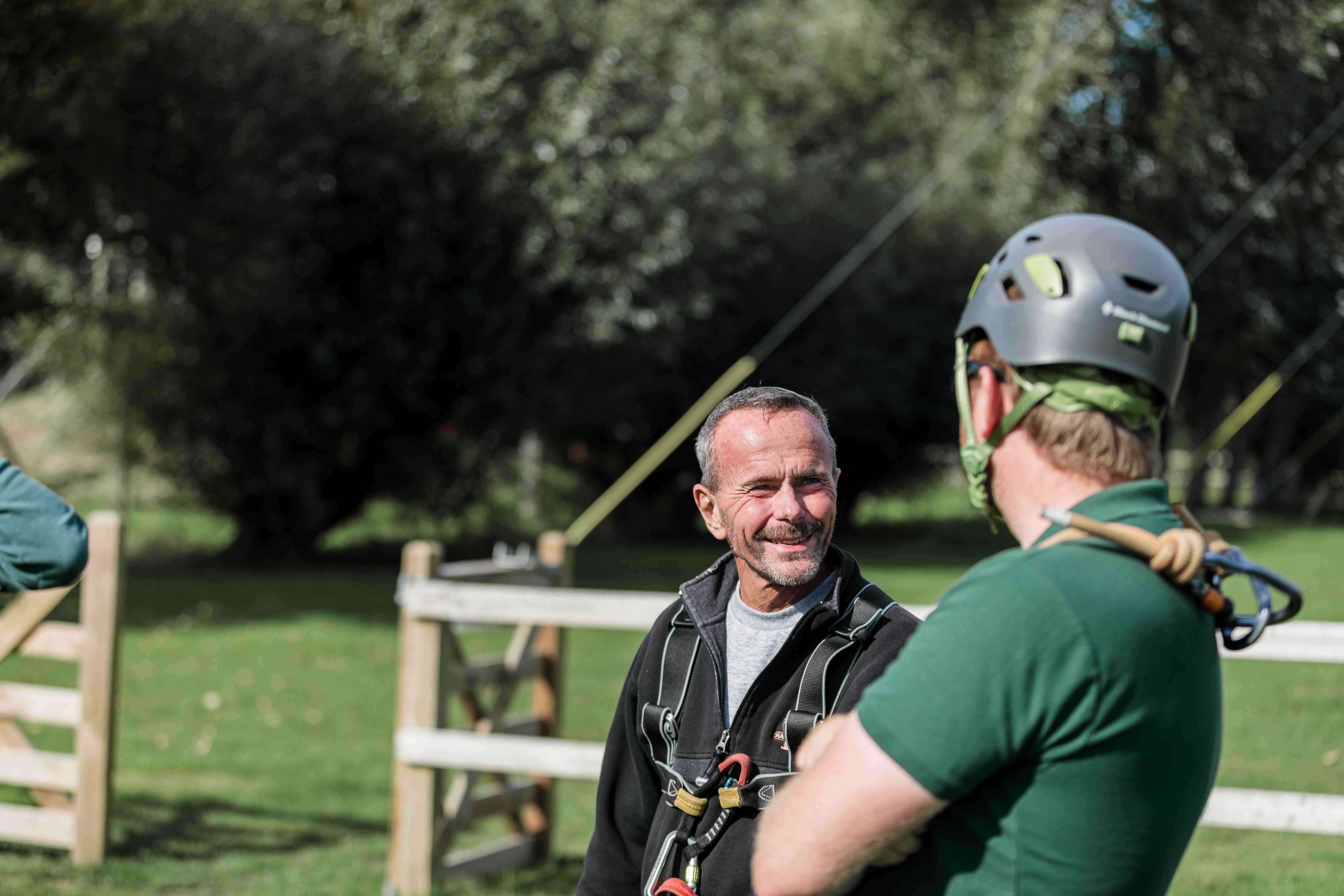 Two men wearing climbing harnesses outdoors, one with a helmet, talking by a wooden fence.