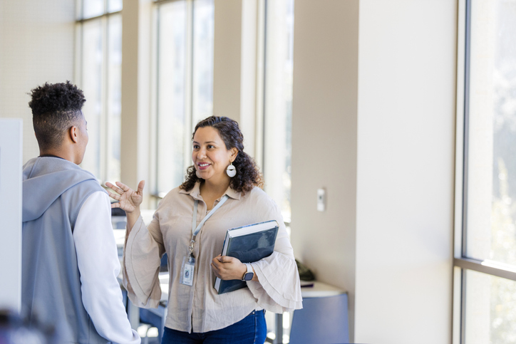 A woman with a lanyard talks to a young man in a bright, modern indoor setting.