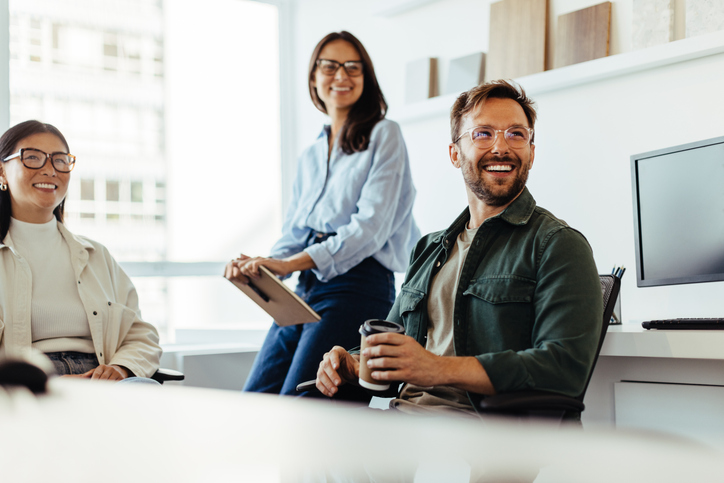 Three smiling colleagues in an office, one holding a coffee cup, others with tablet and sitting.