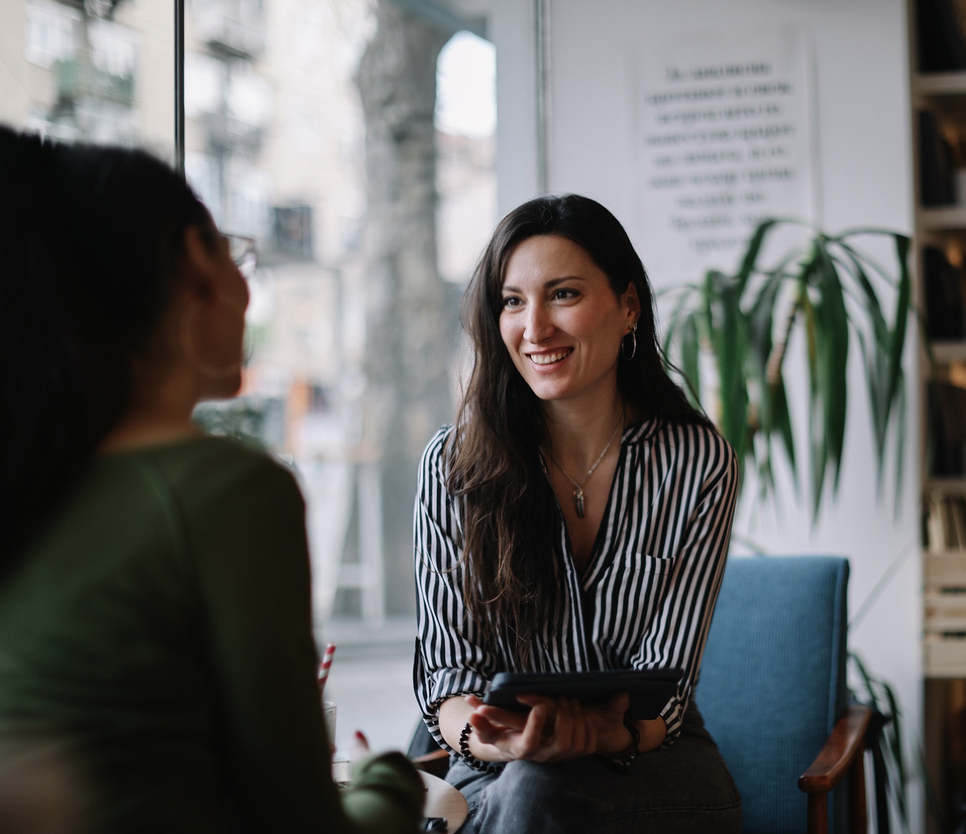 Smiling woman holding tablet while talking to another seated woman in a cozy indoor setting.