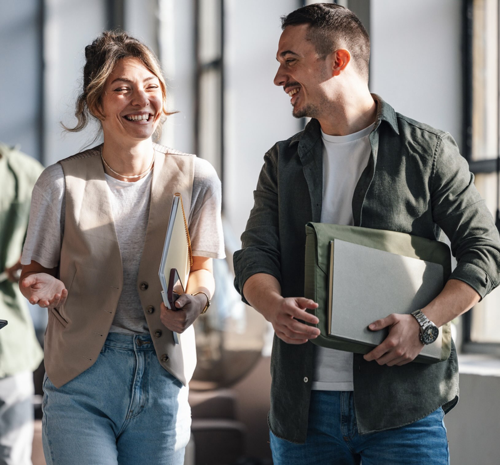 Smiling woman and man walking indoors, holding notebooks and talking happily in natural light.