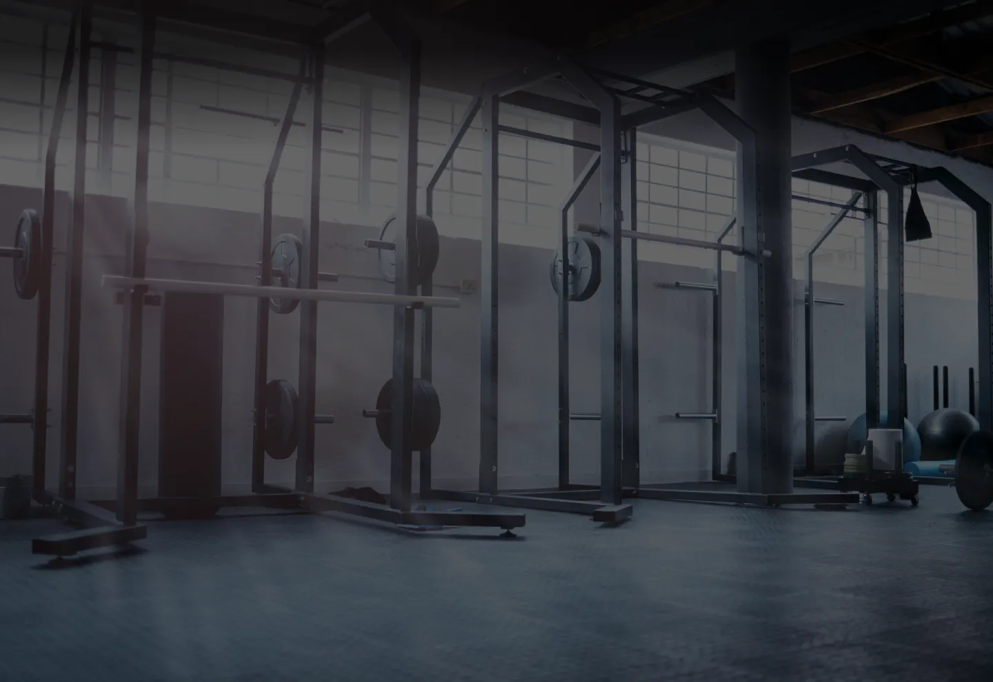 Empty gym with squat racks, barbells, and weight plates next to large windows.