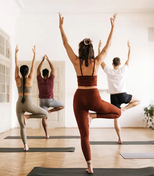 A group of four people practicing yoga indoors, standing on one leg with arms raised in a tree pose on yoga mats.