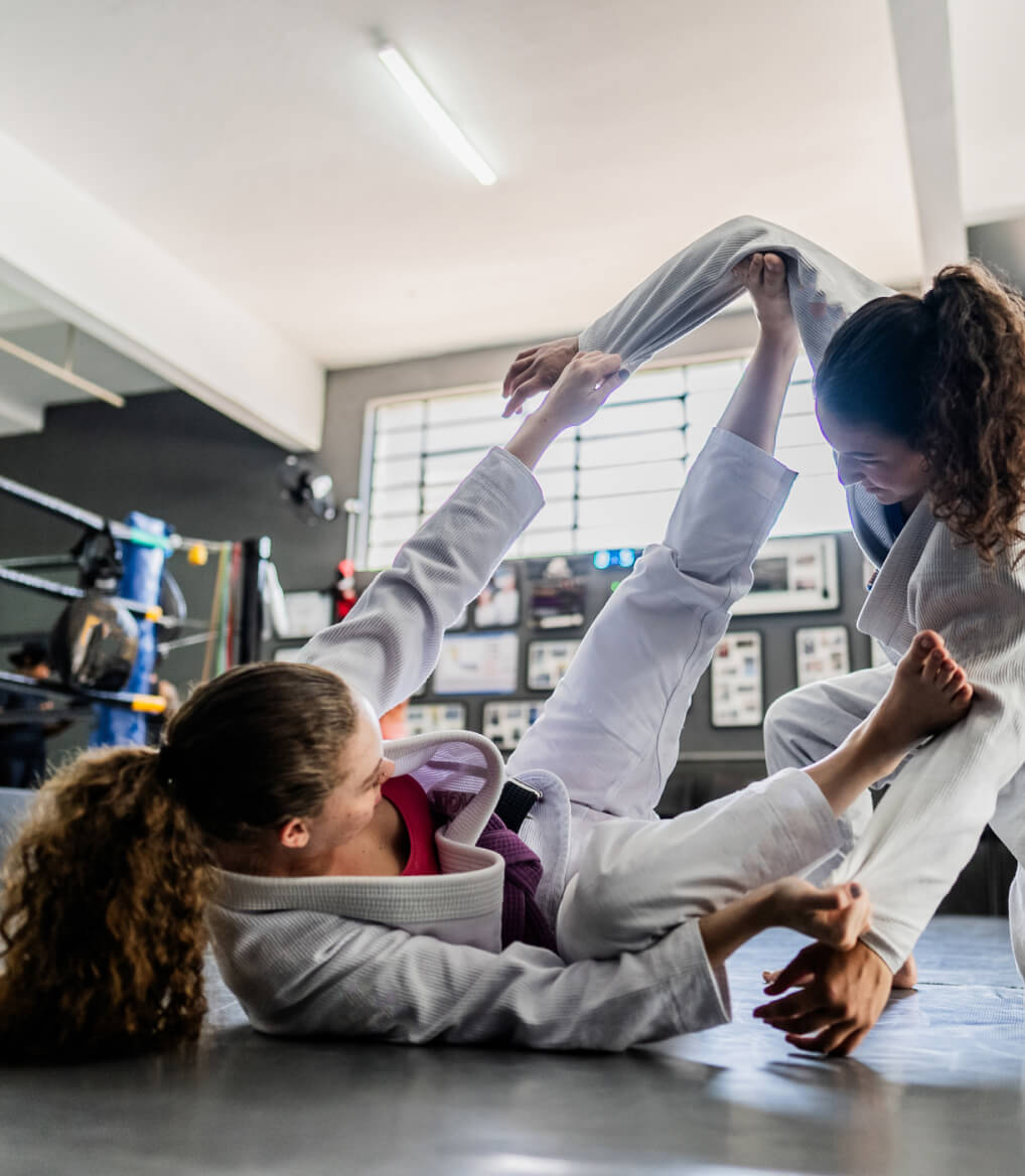 Two women practicing Brazilian Jiu-Jitsu in a gym, wearing white gis and engaging in ground grappling.
