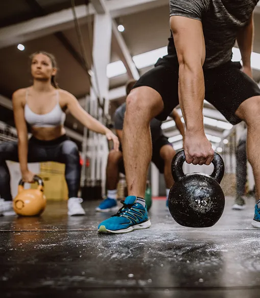 Two people in athletic wear lifting kettlebells in a gym with a focus on their lower bodies and kettlebells.