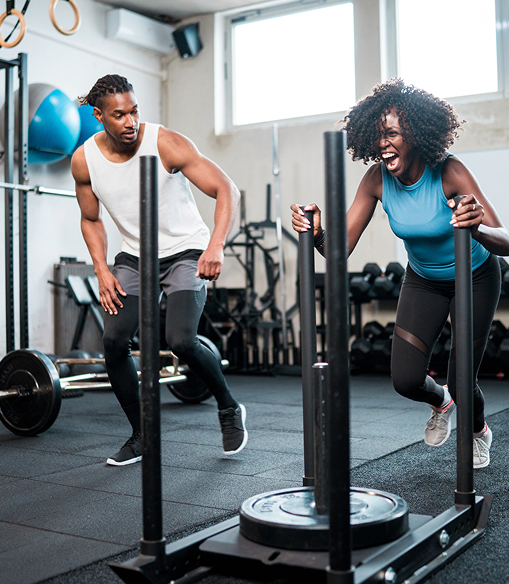 Two people exercising in a gym, one pushing weighted sled and the other running beside it.