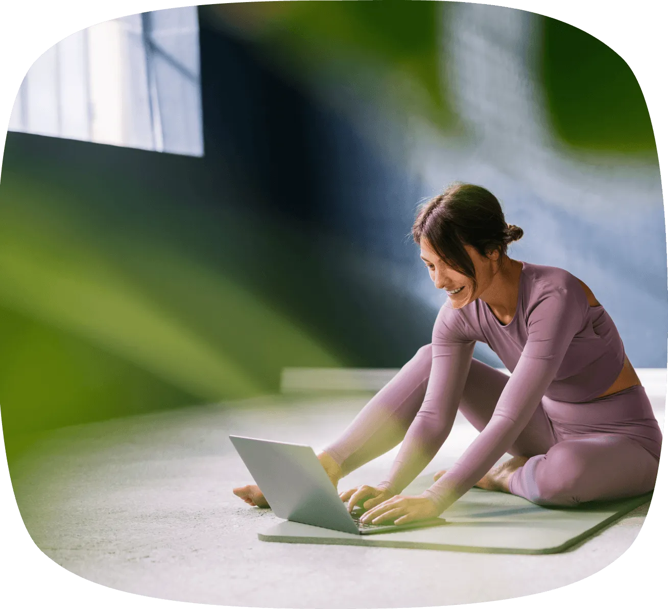 Woman in mauve workout clothes sitting on a yoga mat using a laptop while stretching her legs indoors.