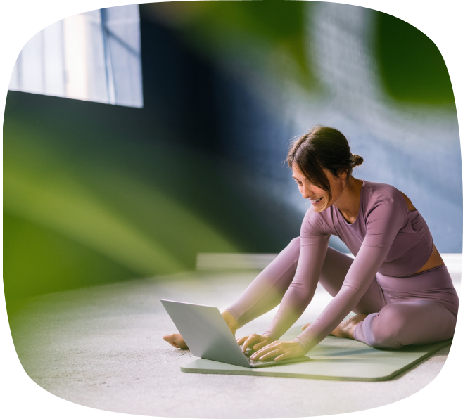 Woman in mauve workout clothes sitting on a yoga mat using a laptop while stretching her legs indoors.