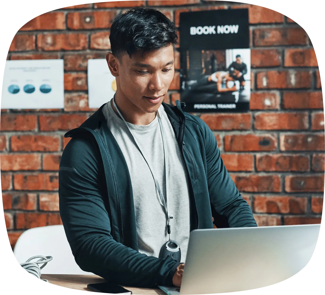 Focused man wearing a whistle around his neck working on a laptop in a gym with a brick wall and fitness posters behind him.