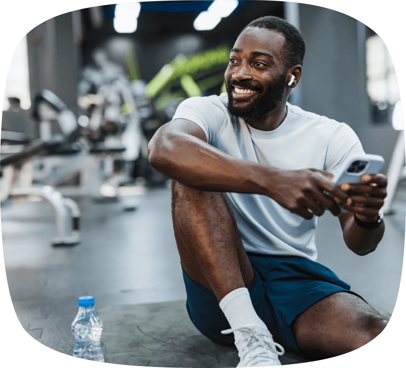 Smiling man in workout clothes sitting on a gym floor holding a smartphone with a water bottle nearby.