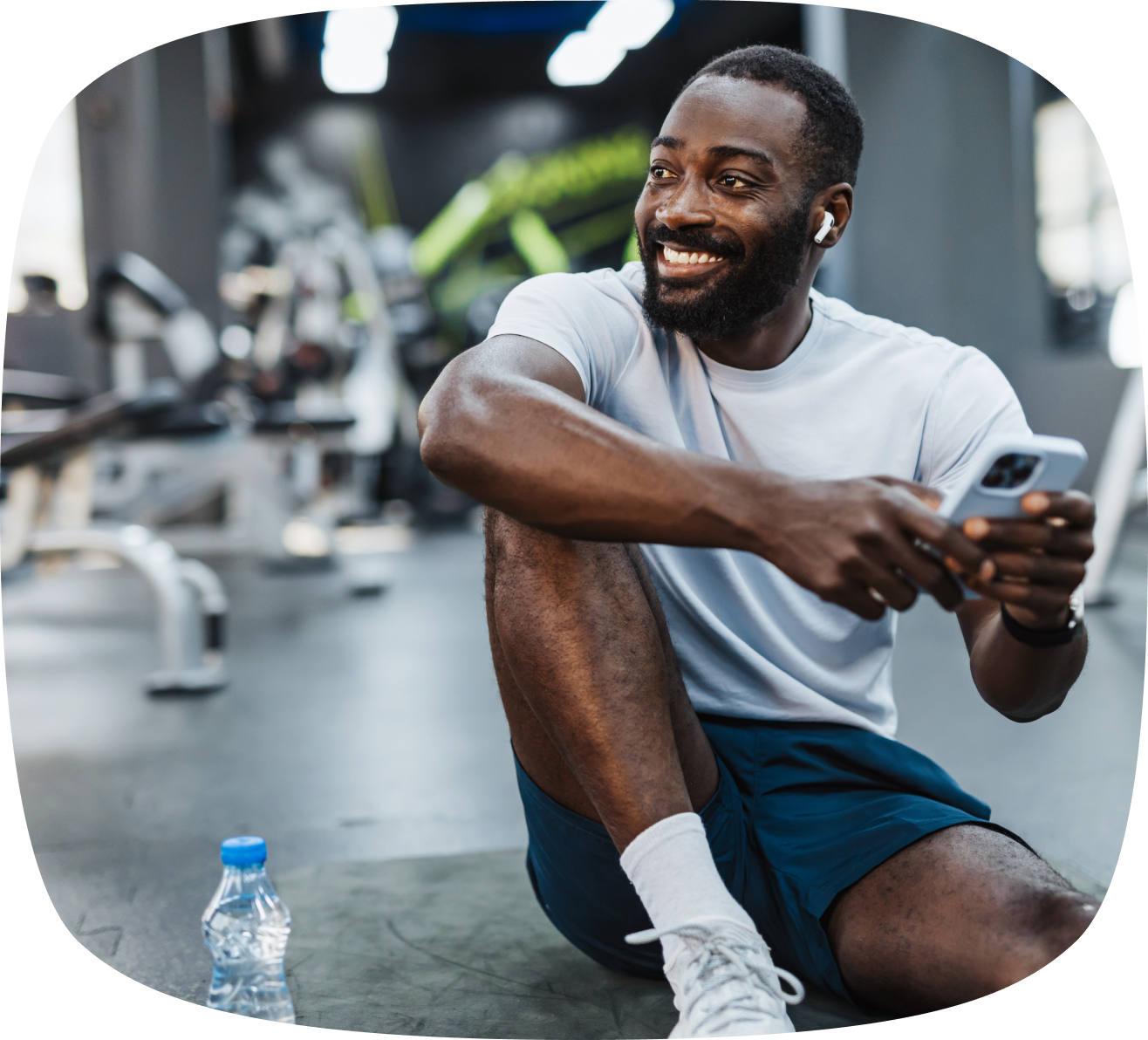 Smiling man in workout clothes sitting on a gym floor holding a smartphone with a water bottle nearby.