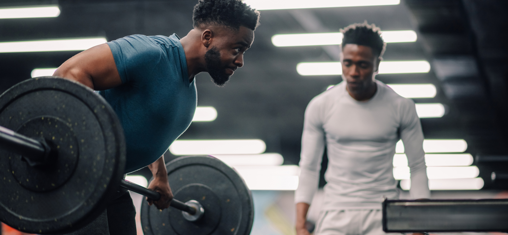 A coach watches an athlete set up a barbell row in a gym