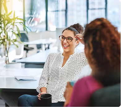 Two women talking and smiling in a bright modern office, one holding a black mug.