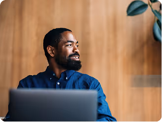 Smiling man with beard looking to the side behind an open laptop in a wooden-paneled room.
