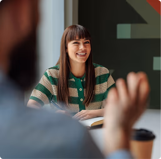Smiling woman with long brown hair wearing a green and white striped shirt, sitting at a table with a coffee cup and notebook, talking to a blurred person in the foreground.