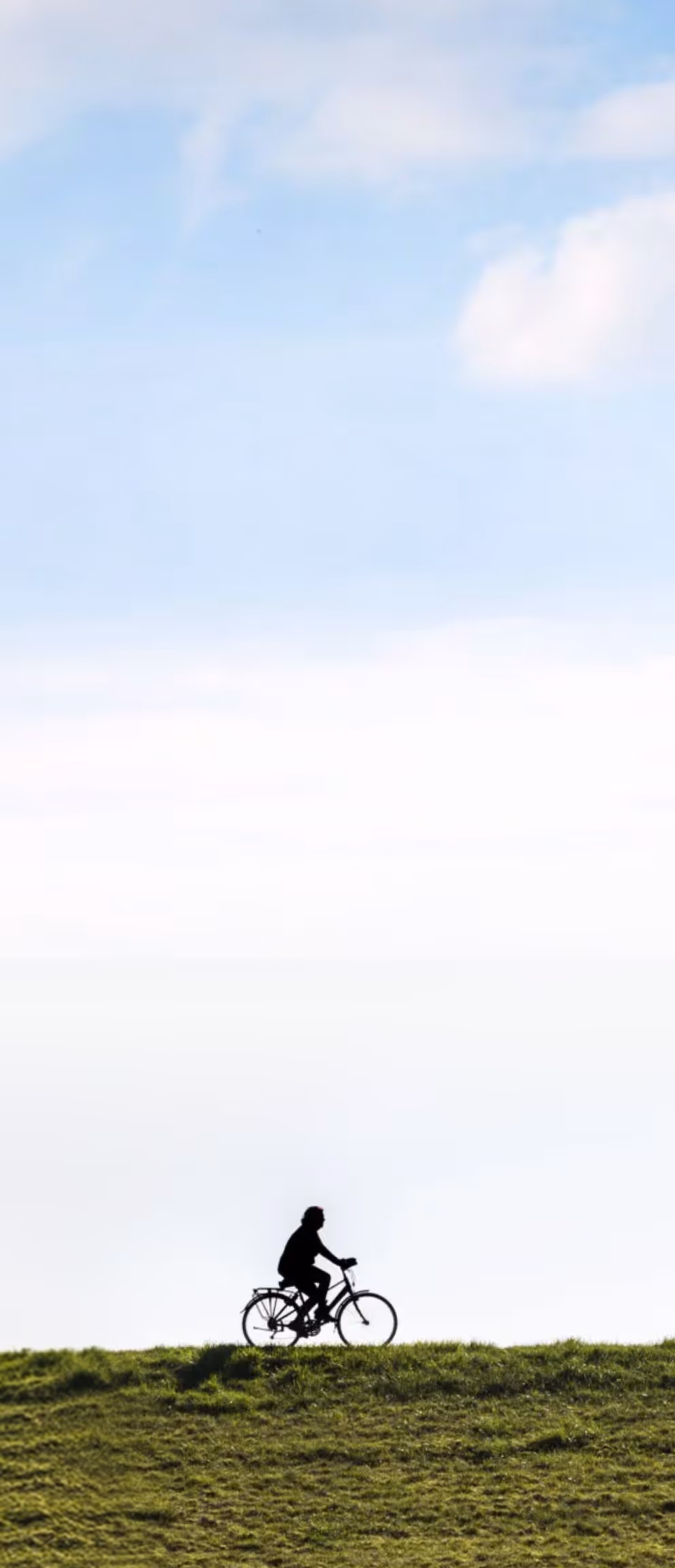 Silhouette of a person riding a bicycle on grass under a blue sky with scattered clouds.
