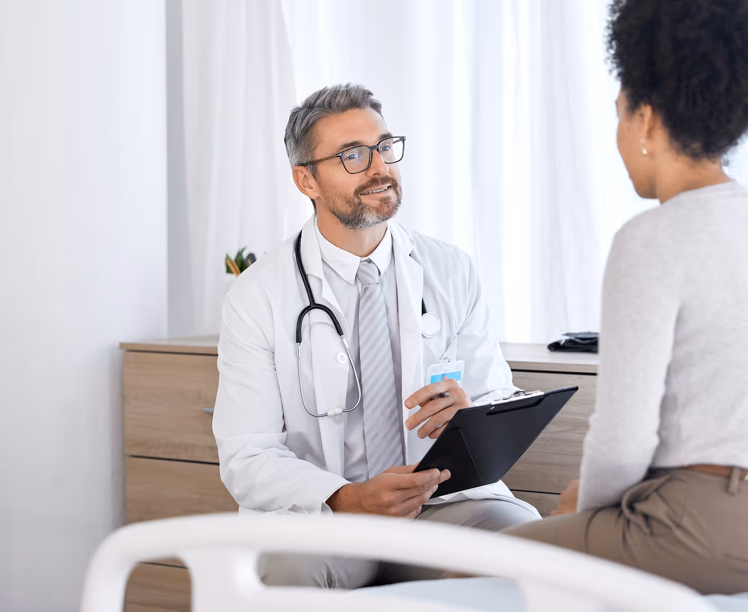 Male doctor with glasses talking to seated female patient while holding a clipboard.