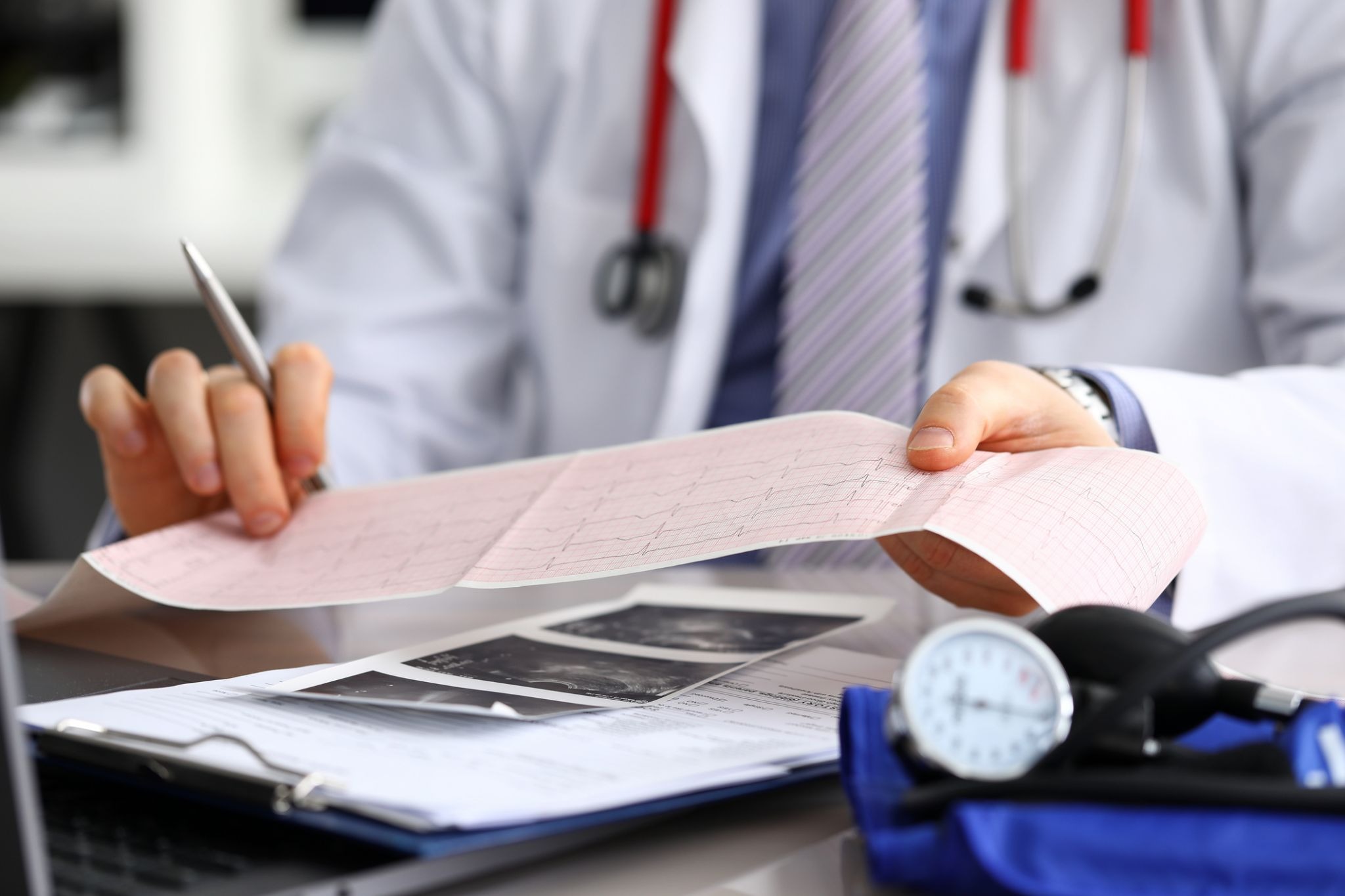 A physician in a white coat and stethoscope reviews a printed ECG tracing at a desk, with a blood pressure cuff and patient records nearby.