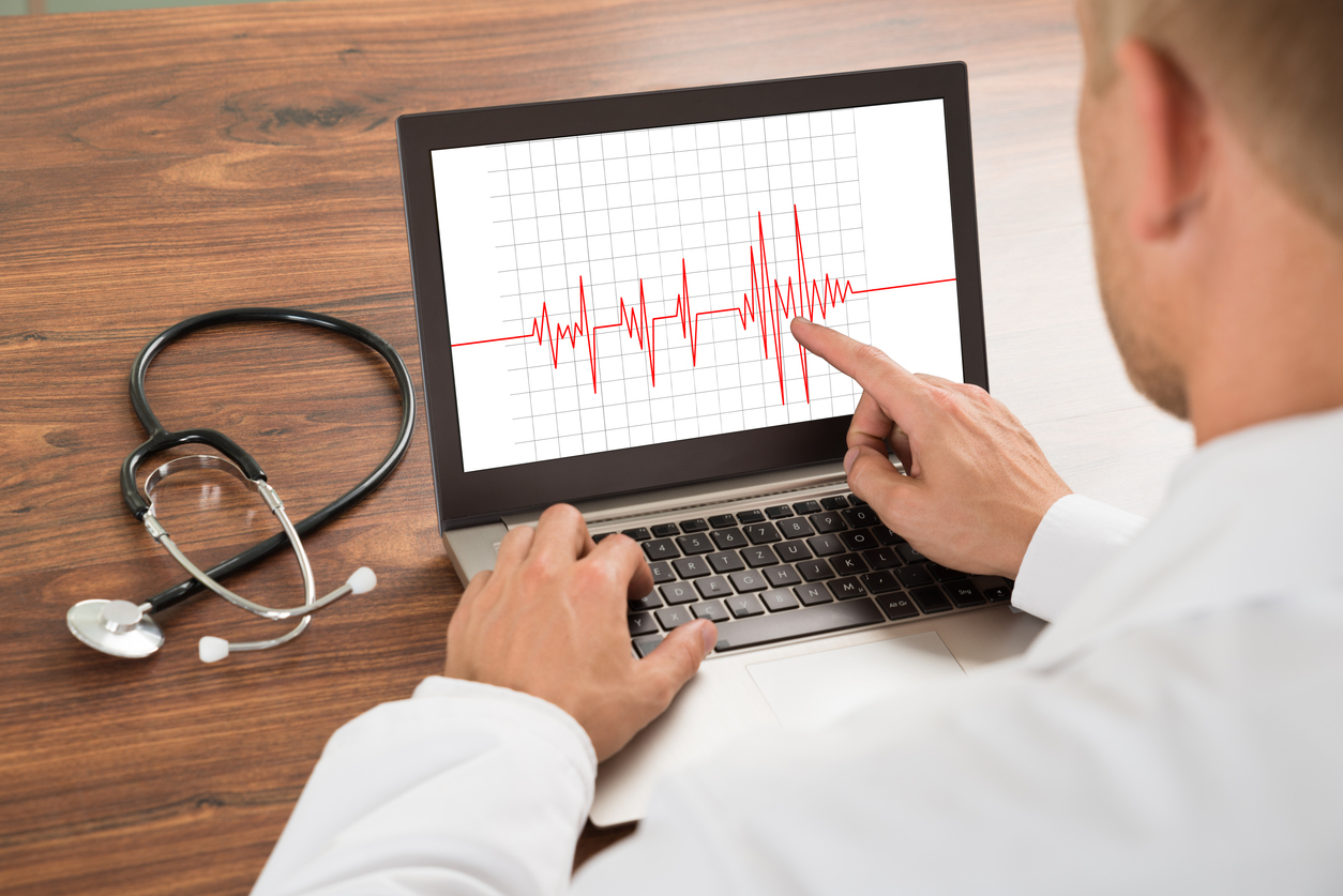 A physician in a white coat points at an ECG tracing on a laptop screen, with a stethoscope resting on the wooden desk beside it.