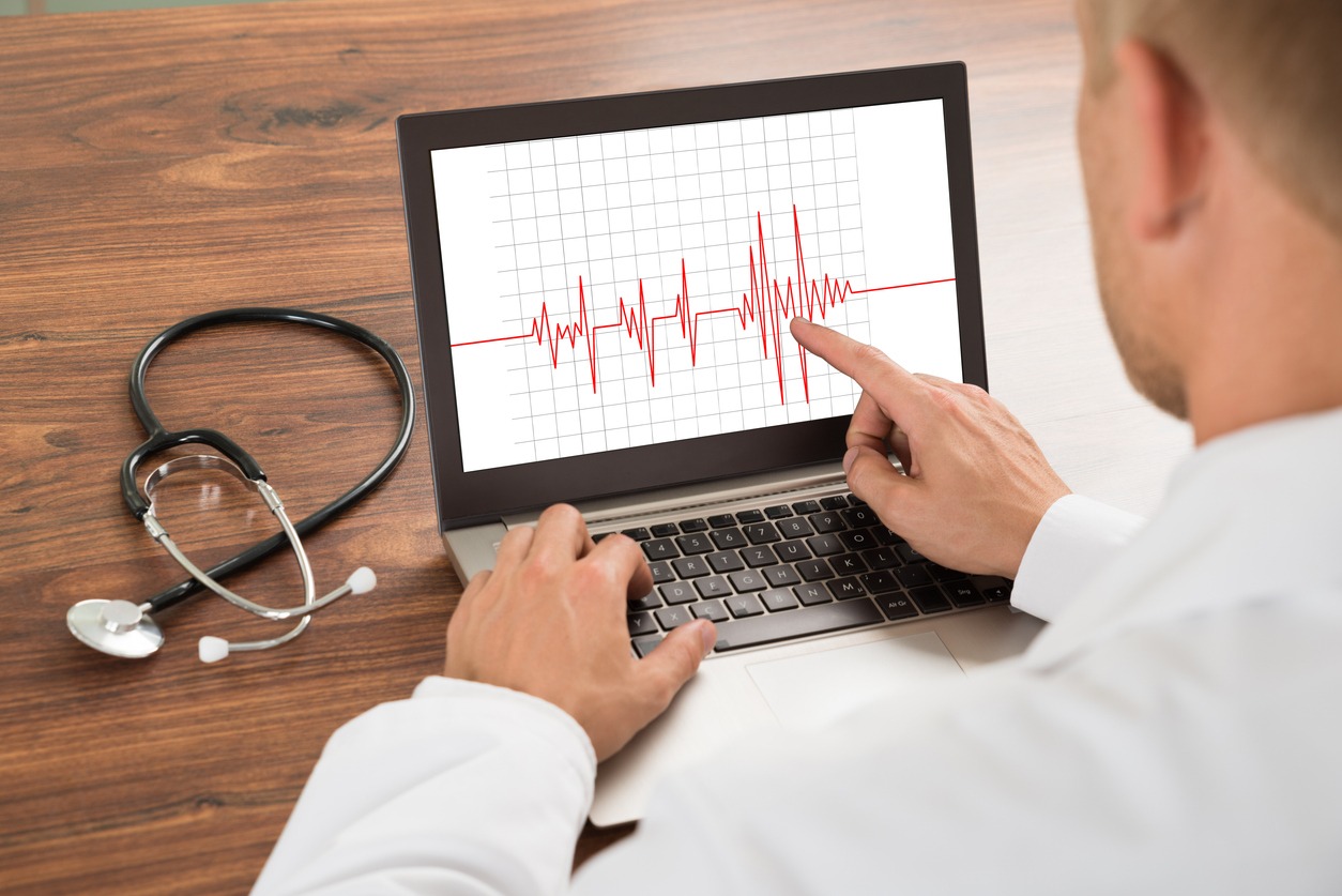 A physician in a white coat points at an ECG tracing on a laptop screen, with a stethoscope resting on the wooden desk beside it.