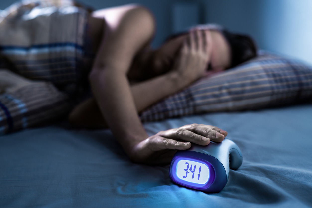 A person lies awake in bed at night, hand resting on an alarm clock reading 3:41, unable to sleep.