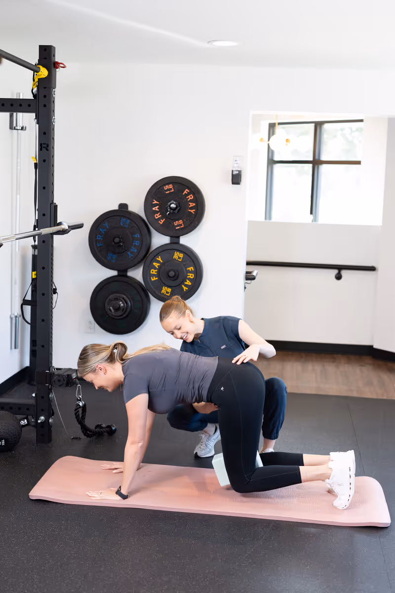 Dr. Ashley instructs a woman performing a quadruped exercise on a pink mat in a gym.