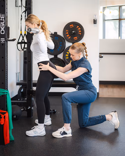 Dr. Ashley in blue assisting woman in white doing a barbell squat in a gym.