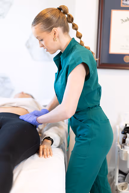 Dr. Ashley in green scrubs with gloves examining a patient's abdomen on a medical bed.