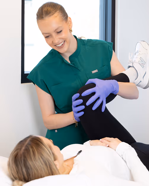 Dr. Ashley in green uniform and purple gloves helping a patient stretch her leg in a clinic.