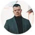 Homme portant un col roulé noir et un blazer gris souriant devant un ciel clair.