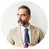 Homme barbu avec costume beige et chemise blanche regardant à droite, souriant légèrement.