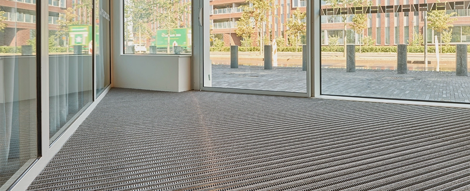 Grey EMCO entrance mat with brush inserts inside a glass entrance facing a plaza