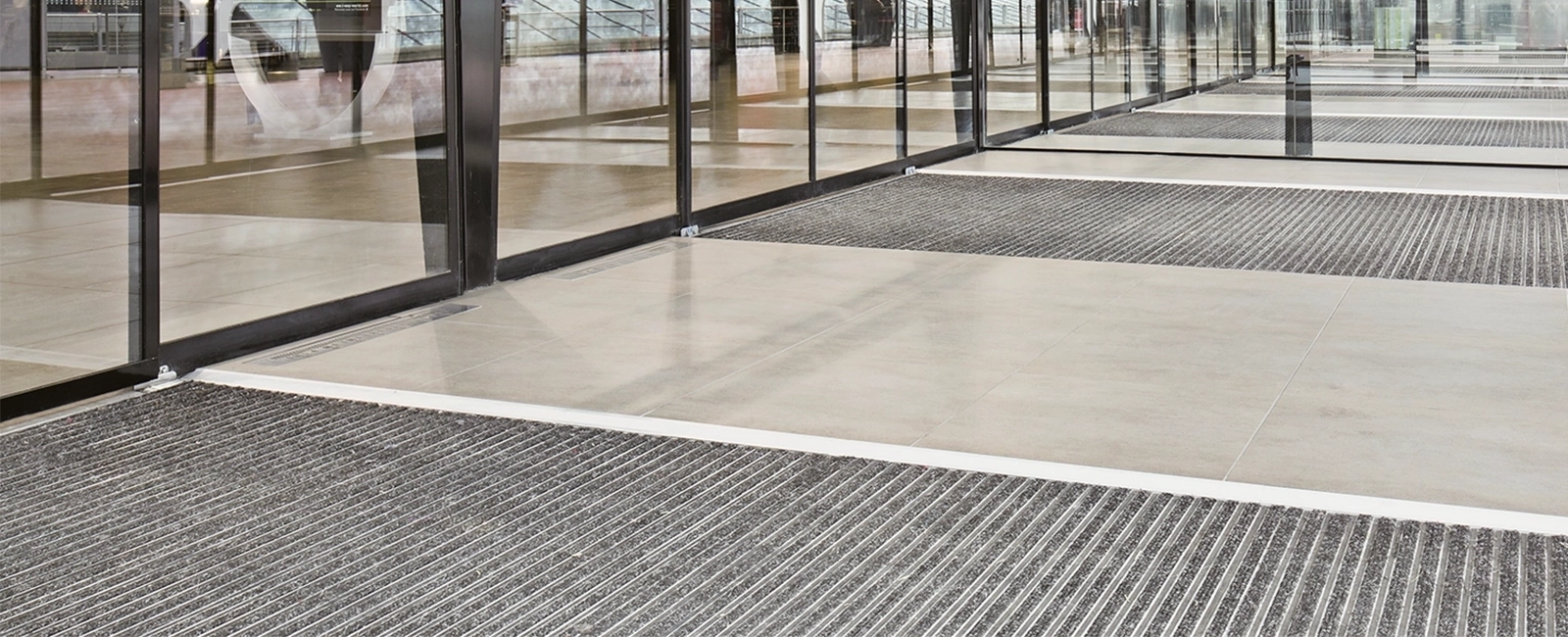 Recessed EMCO entrance mats with grey ribbed inserts in a glass entrance hall