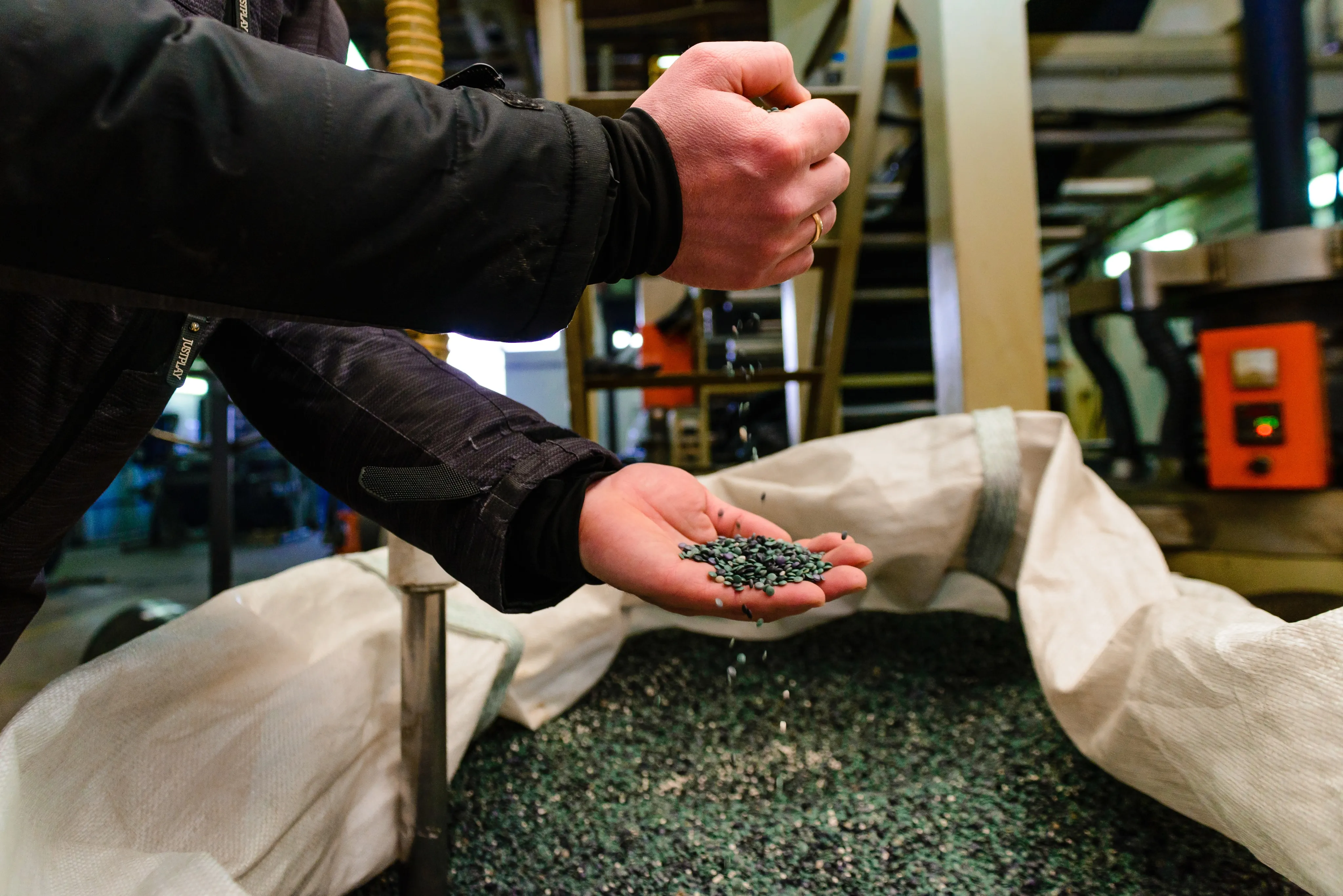 Person holding and sifting dark pellets over a large white sack full of bioplastic pellets in a plant.