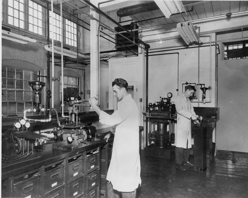 Two men in white lab coats work with scientific instruments in a laboratory with large windows.